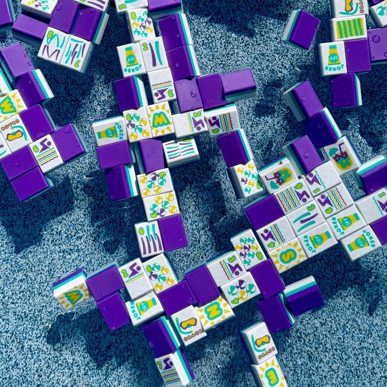 May include: Overhead shot of a collection of Mahjong tiles in white, purple, and teal. The tiles feature various symbols, numbers, and the word "Joker". The tiles are scattered on a textured, light blue surface.