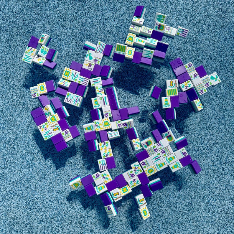 May include: Overhead shot of scattered Mahjong tiles. The tiles are primarily white with colorful symbols and purple edges. The background is a textured, light blue surface.
