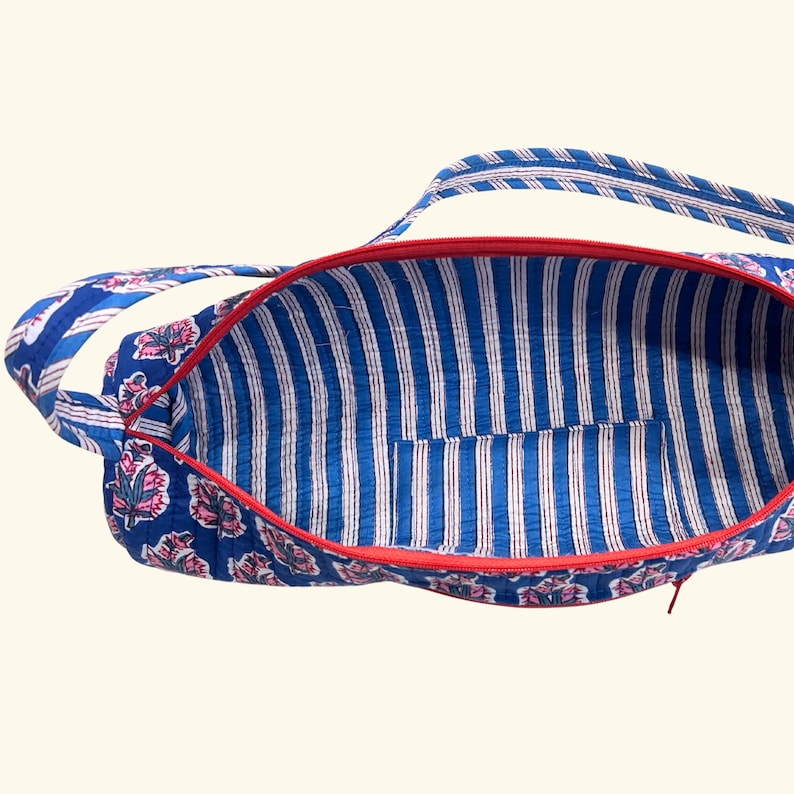 May include: A quilted fabric bag with a blue and white floral exterior pattern and a red zipper closure. The interior features a blue and white vertical striped lining with an open pocket. The bag includes two matching quilted fabric shoulder straps.