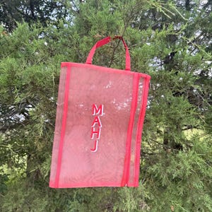 Pu&ograve; includere: Una borsa tote in rete rossa con manico e finiture abbinate. La borsa presenta le lettere ricamate bianche "MAHJ" e una chiusura con cerniera. La borsa &egrave; appesa a un ramo.
