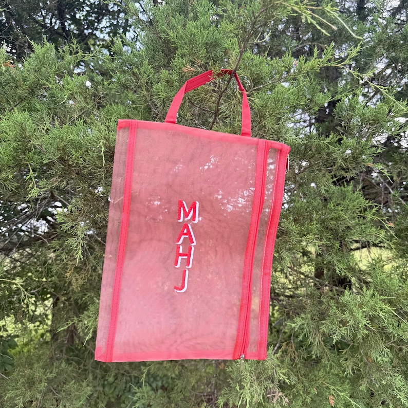 Pu&ograve; includere: Una borsa tote in rete rossa con manico e finiture abbinate. La borsa presenta le lettere ricamate bianche "MAHJ" e una chiusura con cerniera. La borsa &egrave; appesa a un ramo.