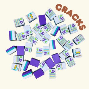 May include: A collection of white, purple, and teal Mahjong tiles scattered on a light beige surface. The tiles feature various colorful designs, including wavy lines, diving masks, and the number 3. The word "CRACKS" is written in brown letters.