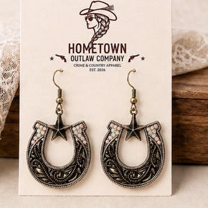 May include: A pair of bronze-toned horseshoe earrings featuring a central five-pointed star and small iridescent accents. The earrings are displayed on a cream-colored card printed with the text HOMETOWN OUTLAW COMPANY CRIME & COUNTRY APPAREL EST. 2026.