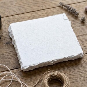 Può includere: Una pila di carta quadrata, bianca e fatta a mano con bordi sfrangiati. Accanto alla carta si trovano un piccolo flacone di inchiostro nero, un pennino in ottone, un rametto di lavanda essiccata, due piccole pietre lisce e un gomitolo di spago.