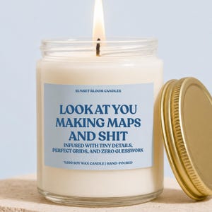 May include: A clear glass jar candle with a light blue label featuring the text SUNSET BLOOM CANDLES, LOOK AT YOU MAKING MAPS AND SHIT, INFUSED WITH TINY DETAILS, PERFECT GRIDS, AND ZERO GUESSWORK, %100 SOY WAX CANDLE | HAND-POURED, with a gold lid.