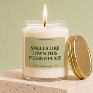 May include: A clear glass jar candle with a lit wick and a sage green label. The label features the text SUNSET BLOOM CANDLES, %100 SOY WAX CANDLE | HAND-POURED, and the phrase SMELLS LIKE I OWN THIS F*CKING PLACE. A gold metal lid sits beside the jar.