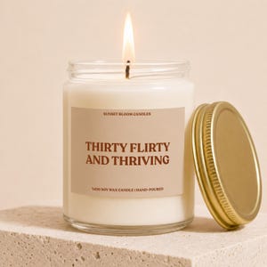 Peut inclure: Une bougie en pot de verre transparent avec une mèche allumée et un couvercle en métal doré posé à côté. L'étiquette porte le texte SUNSET BLOOM CANDLES, THIRTY FLIRTY AND THRIVING, et %100 SOY WAX CANDLE | HAND-POURED en lettres marron foncé.