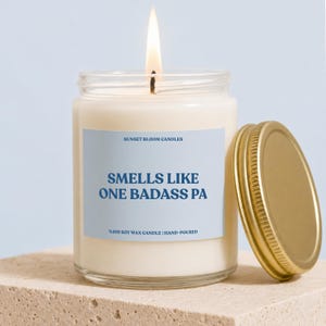 May include: A clear glass jar candle with a lit wick and white wax. The label is light blue with dark blue text reading SUNSET BLOOM CANDLES, SMELLS LIKE ONE BADASS PA, and %100 SOY WAX CANDLE | HAND-POURED. A gold metal lid rests beside the jar.