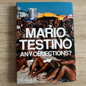 Può includere: Un libro con copertina rigida intitolato Mario Testino Any Objections? pubblicato da Phaidon. La copertina presenta una vivace scena di spiaggia baciata dal sole con numerose persone che si rilassano sulla sabbia sotto ombrelloni gialli e rossi, con edifici cittadini sullo sfondo.