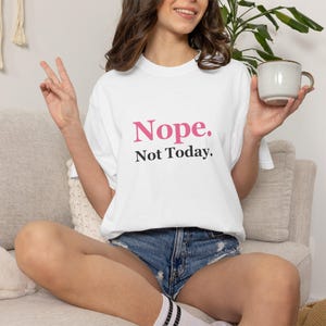 Puede incluir: Una camiseta blanca de manga corta con el texto "Nope." en letras rosas sobre el texto "Not Today." en letras negras. La camiseta tiene un cuello redondo y un corte estándar, ofreciendo un aspecto limpio y sencillo para uso diario.