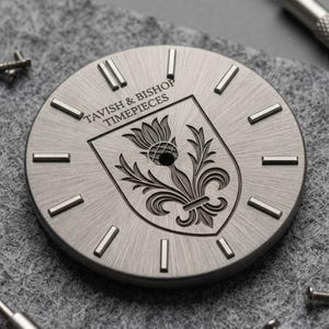 May include: A circular, brushed silver watch dial featuring raised rectangular hour markers. The center displays an engraved shield with a thistle and fleur-de-lis motif. The text TAVISH & BISHOP TIMEPIECES is printed in black above the shield.