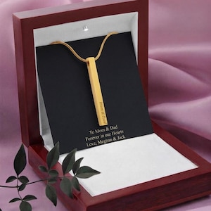 Puede incluir: Un collar con colgante de barra vertical en tono dorado sobre una tarjeta negra dentro de una caja de madera color caoba. La barra está grabada con el texto "Mom & Dad" y la tarjeta presenta el texto "To Mom & Dad Forever in our Hearts Love, Meghan & Jack".
