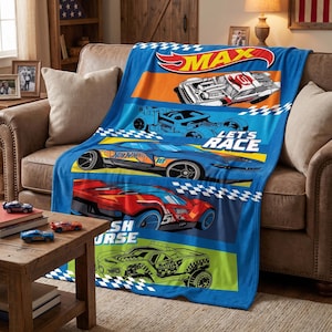 May include: A blue fleece throw blanket featuring Hot Wheels race cars in red, orange, blue, and green. The design includes the Hot Wheels logo, the text MAX and LET'S RACE, and checkered flag patterns across the fabric surface.