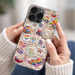 May include: A smartphone case featuring a colorful craft-themed pattern with embroidery hoops, yarn baskets, scissors, needles, and floral cross-stitch designs. The name Violet is printed in a brown script font in the center of the case.