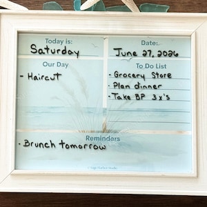 Peut inclure: Un tableau effaçable à sec dans un cadre blanc avec un fond sur le thème de la plage. Il comporte des sections pour Today is: Saturday, Date: June 27, 2026, Our Day, To Do List et Reminders. Le texte manuscrit indique Haircut, Grocery Store, Plan dinner.