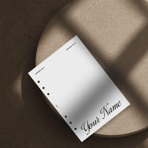 May include: A white rectangular planner insert page with six circular binder holes along the left edge. The page features the printed text "PERSONAL TEXT 1" and "PERSONAL TEXT 2" at the top, with the script text "Your Name" in the bottom right corner.