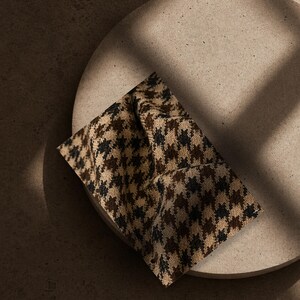 May include: A square piece of fabric featuring a brown, black, and beige houndstooth pattern. The textile has a visible woven texture and is draped with soft folds, creating a dimensional appearance with light and shadow across the surface.