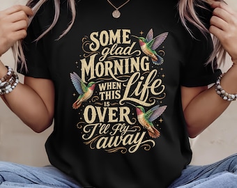 以下が含まれることがあります： 3羽のカラフルなハチドリと「Some glad MORNING WHEN THIS IS OVER I'll fly away」というテキストがプリントされた黒いTシャツ。デザインには、装飾的な曲線と小さな星のアクセントが施されたゴールドとクリーム色の文字が使用されています。