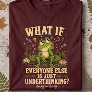 May include: A maroon t-shirt featuring a graphic of a green frog sitting on a lily pad. The text reads: WHAT IF EVERYONE ELSE IS JUST UNDERTHINKING? Asking For A Frog. The design includes cattails, dragonflies, water lilies, and light bulbs.