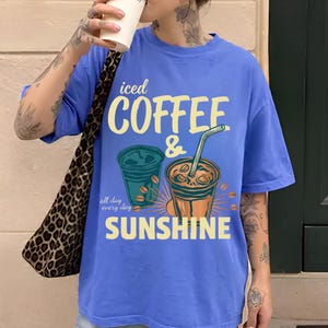 以下が含まれることがあります： 黄色と白で「iced COFFEE & SUNSHINE all day every day」という文字がプリントされた、ペリウィンクルブルーの半袖Tシャツ。デザインには、ストロー付きのアイスコーヒーカップ2つと、散らばったコーヒー豆のイラストが描かれています。