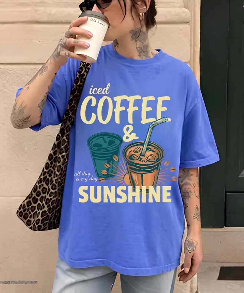 以下が含まれることがあります： 黄色と白で「iced COFFEE & SUNSHINE all day every day」という文字がプリントされた、ペリウィンクルブルーの半袖Tシャツ。デザインには、ストロー付きのアイスコーヒーカップ2つと、散らばったコーヒー豆のイラストが描かれています。