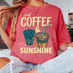 以下が含まれることがあります： 「iced COFFEE & SUNSHINE all day every day」というテキストがプリントされたコーラルカラーのTシャツ。デザインには、ストロー付きのカップを含むアイスコーヒーが入った2つのプラスチックカップのイラストと、散らばったコーヒー豆が描かれています。