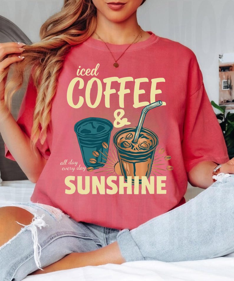 以下が含まれることがあります： 「iced COFFEE & SUNSHINE all day every day」というテキストがプリントされたコーラルカラーのTシャツ。デザインには、ストロー付きのカップを含むアイスコーヒーが入った2つのプラスチックカップのイラストと、散らばったコーヒー豆が描かれています。