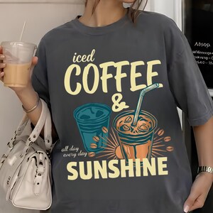 以下が含まれることがあります： 「iced COFFEE & SUNSHINE all day every day」というテキストがプリントされたチャコールグレーの半袖Tシャツ。デザインには、ストロー付きのアイスコーヒーカップ2つと、黄色と青で描かれたコーヒー豆のイラストが含まれています。