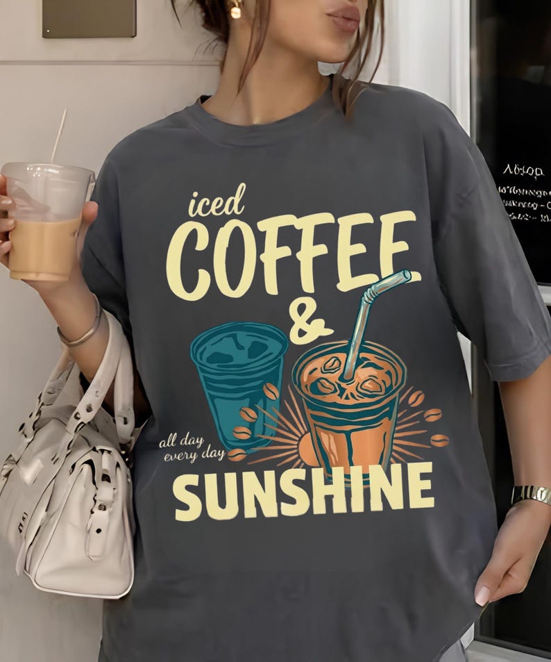 以下が含まれることがあります： 「iced COFFEE & SUNSHINE all day every day」というテキストがプリントされたチャコールグレーの半袖Tシャツ。デザインには、ストロー付きのアイスコーヒーカップ2つと、黄色と青で描かれたコーヒー豆のイラストが含まれています。