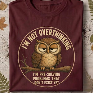 Könnte beinhalten: Ein kastanienbraunes T-Shirt mit einer kreisförmigen Grafik einer braunen Eule, die mit halb geschlossenen Augen auf einem Ast sitzt. Der Text lautet: I'M NOT OVERTHINKING I'M PRE-SOLVING PROBLEMS THAT DON'T EXIST YET. Das Design ist hellbeige.