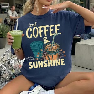 以下が含まれることがあります： 「iced COFFEE & SUNSHINE」および「all day every day」というテキストがプリントされた、ネイビーブルーの半袖Tシャツ。デザインには、ストロー付きのアイスコーヒーカップ2つと、黄色、オレンジ、青緑色のコーヒー豆のイラストが描かれています。