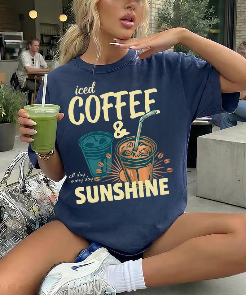 以下が含まれることがあります： 「iced COFFEE & SUNSHINE」および「all day every day」というテキストがプリントされた、ネイビーブルーの半袖Tシャツ。デザインには、ストロー付きのアイスコーヒーカップ2つと、黄色、オレンジ、青緑色のコーヒー豆のイラストが描かれています。
