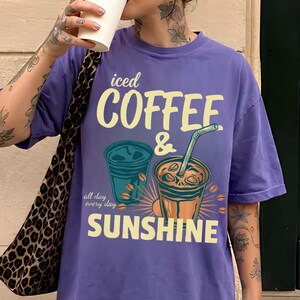 以下が含まれることがあります： 「iced COFFEE & SUNSHINE」および「all day every day」というテキストがプリントされた紫色の半袖Tシャツ。デザインには、ストロー付きのアイスコーヒーカップ2つと、黄色と青色のコーヒー豆のイラストが描かれています。