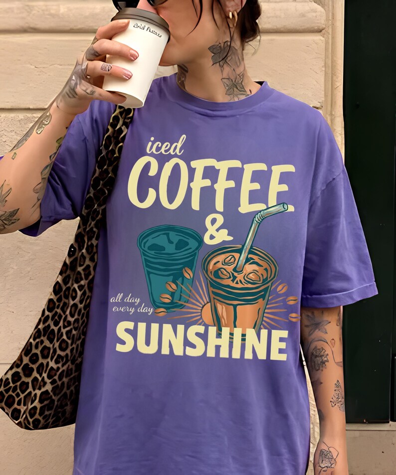 以下が含まれることがあります： 「iced COFFEE & SUNSHINE」および「all day every day」というテキストがプリントされた紫色の半袖Tシャツ。デザインには、ストロー付きのアイスコーヒーカップ2つと、黄色と青色のコーヒー豆のイラストが描かれています。