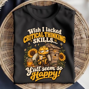 Op de afbeelding: Een zwart t-shirt met een afbeelding van een vermoeide bij die een mok vasthoudt naast een bijenkorf, zonnebloemen en een pot honing. De tekst luidt: Wish I lacked CRITICAL THINKING SKILLS... Y'all seem so Happy! Ontwerp in geel, oranje en wit.