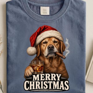 Op de afbeelding: Een blauw t-shirt met ronde hals en een afbeelding van een bruine hond met een rood-witte kerstmuts, die een glas met amberkleurige vloeistof en ijs vasthoudt en een sigaar rookt, met de tekst MERRY CHRISTMAS in het wit onder de hond.