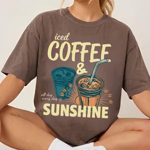 以下が含まれることがあります： ストローとコーヒー豆が描かれた2つのアイスコーヒーのグラフィックがプリントされた茶色の半袖Tシャツ。「iced COFFEE & SUNSHINE」という文字が大きな黄色いフォントで書かれており、左側には「all day every day」という小さな筆記体のテキストが添えられています。