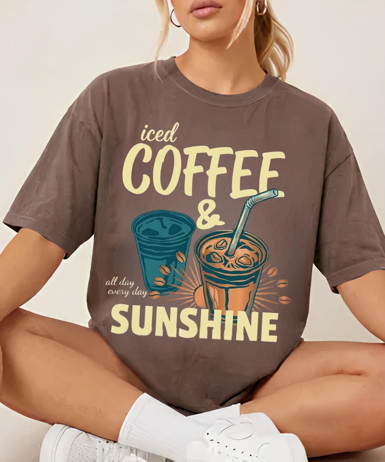 以下が含まれることがあります： ストローとコーヒー豆が描かれた2つのアイスコーヒーのグラフィックがプリントされた茶色の半袖Tシャツ。「iced COFFEE & SUNSHINE」という文字が大きな黄色いフォントで書かれており、左側には「all day every day」という小さな筆記体のテキストが添えられています。