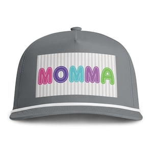 May include: A grey baseball cap featuring a white rectangular patch with thin vertical grey stripes. The word MOMMA is printed in large, colourful, bubble-style letters with a stitched outline effect in pink, purple, blue, purple, and green.