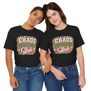 Può includere: Una t-shirt nera con un design grafico caratterizzato da un bordo a scacchi, stelle rosa, un fulmine, una tazzina da caffè e una faccina sorridente. Il testo recita "CHAOS COORDINATOR Club" in un mix di lettere bianche, verde oliva e rosa.