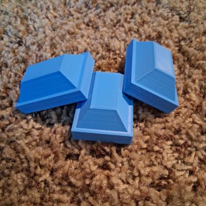 Pode incluir: Três objetos tridimensionais retangulares de cor azul claro com uma parte superior escalonada em forma de pirâmide. Cada peça apresenta uma base plana e uma estrutura superior inclinada de vários níveis com linhas de camada horizontais visíveis.