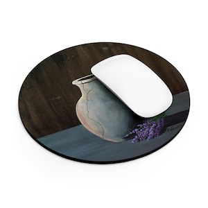 May include: A round mouse pad featuring a printed illustration of a cracked, light-colored ceramic vase resting on a dark surface next to a sprig of purple lavender. A smooth, white computer mouse is positioned on top of the vase illustration.