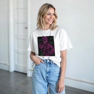 Peut inclure: Un t-shirt blanc à manches courtes avec un motif carré noir central présentant un damier magenta représentant une créature aux grands yeux et aux mains levées, accompagné de petits encadrés de texte vert sur les côtés du motif.