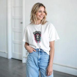 以下が含まれることがあります： ピンクの野球帽とチェーンネックレスを身に着けたエイリアンのグラフィックプリントが施された白い半袖Tシャツ。エイリアンの上のテキストには「THE GOVERNMENT HID THE VIBE」と書かれており、その下にはピンクと青のカラフルなペイント風のストライプが描かれています。