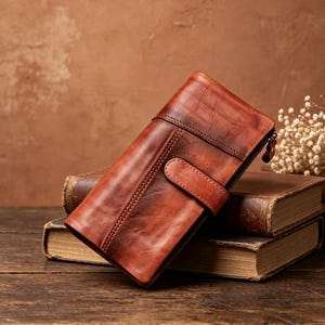 May include: A rectangular, bi-fold wallet in a rich, burnished brown leather with a smooth, polished finish. The wallet features a prominent horizontal strap with a snap closure, visible stitching along the edges, and a side zipper pull.