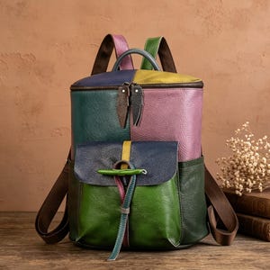Könnte beinhalten: Ein mehrfarbiger Lederrucksack mit Einsätzen in Blau, Grün, Gelb und Violett. Die Tasche verfügt über einen Tragegriff, zwei Schultergurte, ein Hauptfach mit Reißverschluss und zwei Zippern sowie eine Vordertasche mit Knebelverschluss.