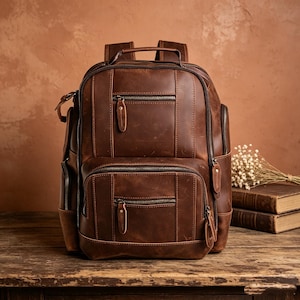 May include: A brown leather backpack featuring multiple zippered compartments, a top handle, and adjustable shoulder straps. The exterior displays a distressed finish with visible stitching, metal zippers, and leather pull tabs on each pocket.