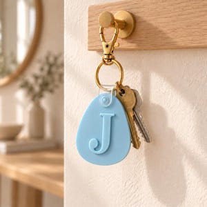 May include: A light blue, egg-shaped keychain featuring a raised letter J in the center. The keychain is attached to a gold-toned metal lobster clasp and a split ring, which holds two silver-toned metal keys. The keychain hangs from a hook.