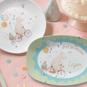 May include: A round white plate and an oval light green plate featuring a cartoon bear riding a bicycle with balloons and gifts. The oval plate includes the text 2nd Birthday and the name Logan in a script font, set against a party theme.