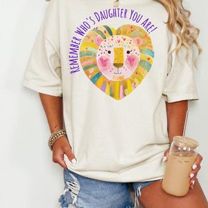 May include: An off-white short-sleeve t-shirt featuring a colorful heart-shaped lion illustration and the purple text "REMEMBER WHO'S DAUGHTER YOU ARE!" printed on the front. The design includes shades of yellow, pink, orange, and green.